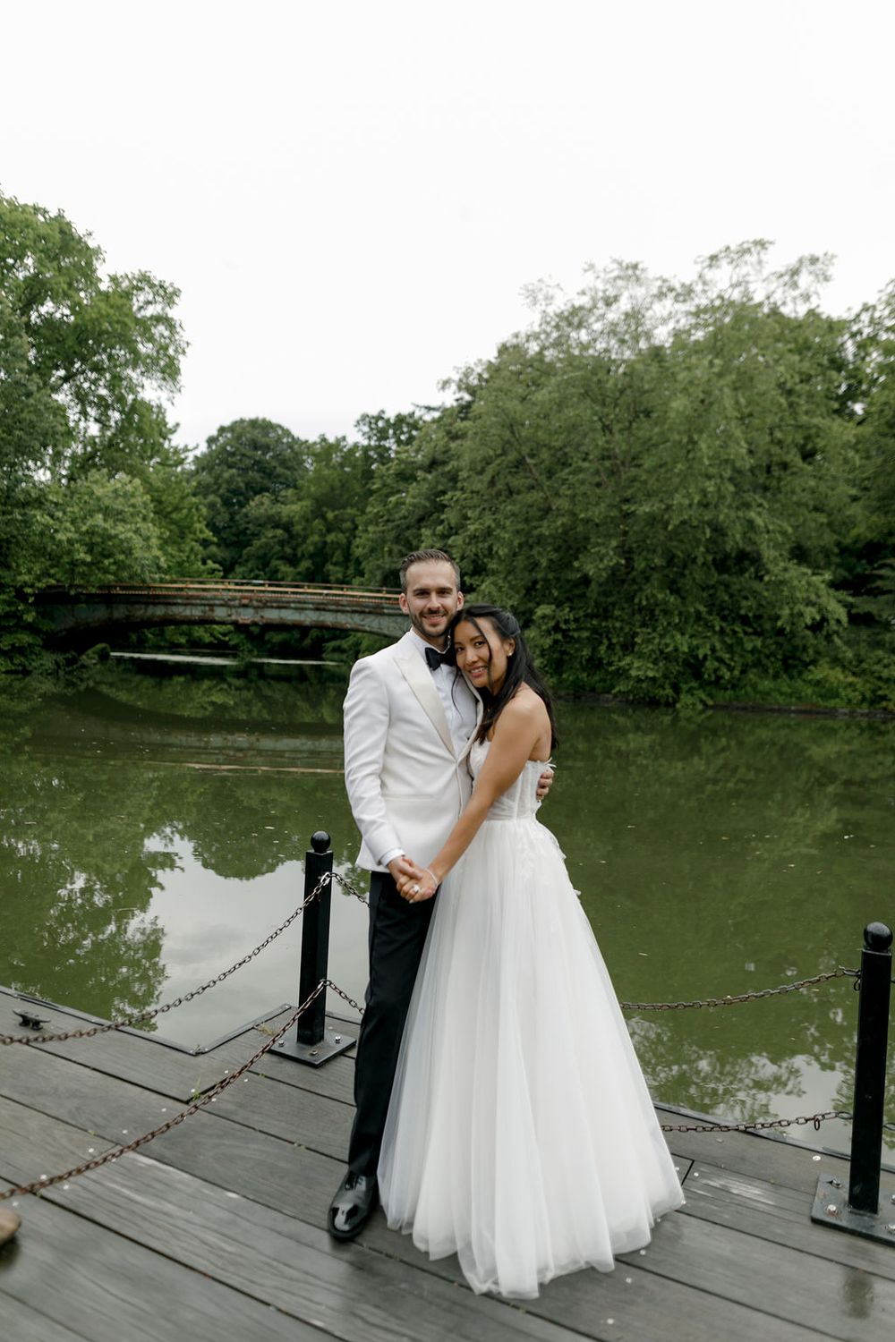 Wedding couple embracing on wooden deck overlooking pond with arched bridge in background.