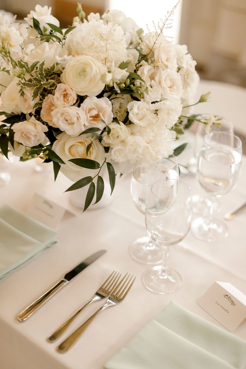 Elegant white floral centerpiece with roses and peonies on formal table setting with wine glasses and silverware.