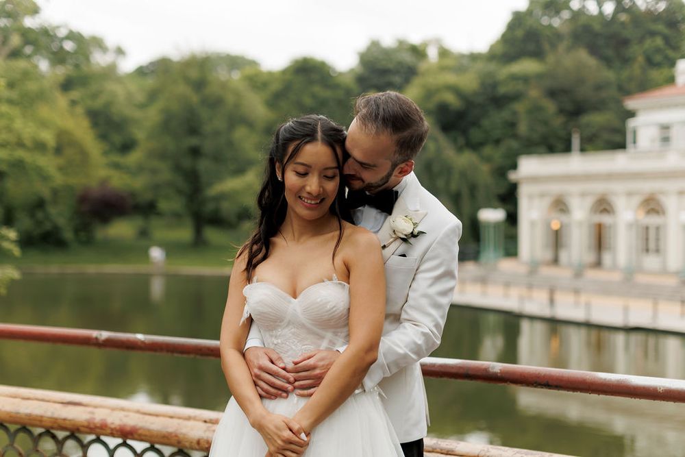 Wedding couple embraces on balcony overlooking scenic lake and classical architecture.