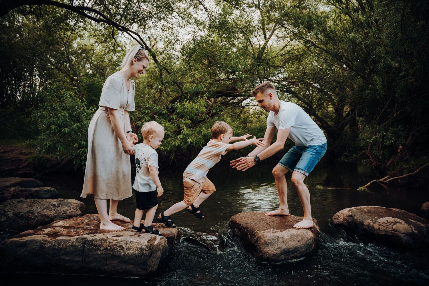 Eine Familien steht barfuß auf Steinen in einem Fluss. Es ist Sommer und alle sind sommerlich gekleidet. Eines der Kinder springt zum Papa.