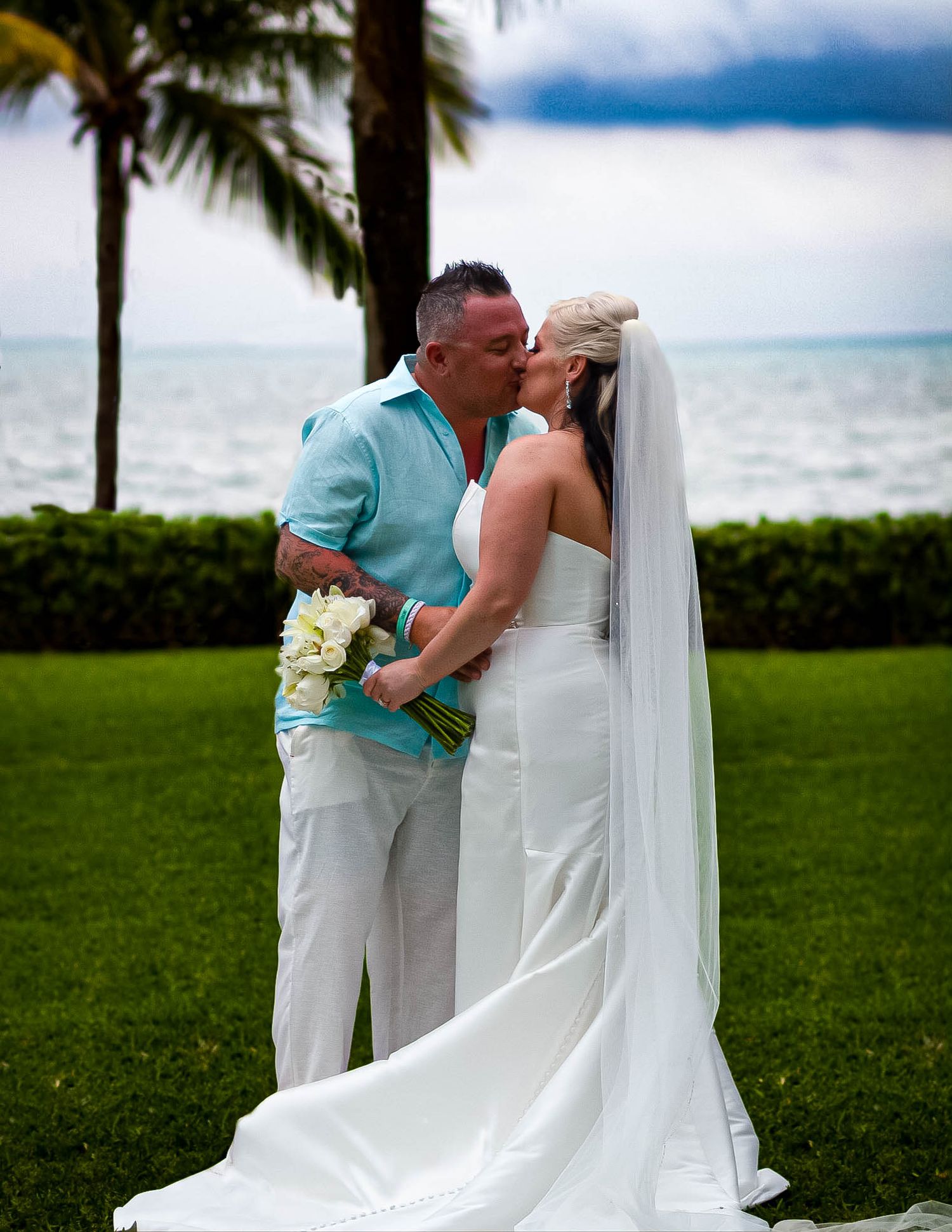 A romantic beach wedding kiss with palm trees and ocean waves in the background.