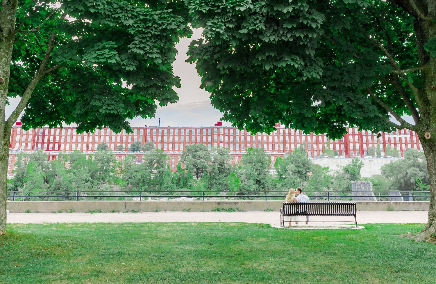 Empty park bench sits in front of a long brick building with manicured lawn and trees.