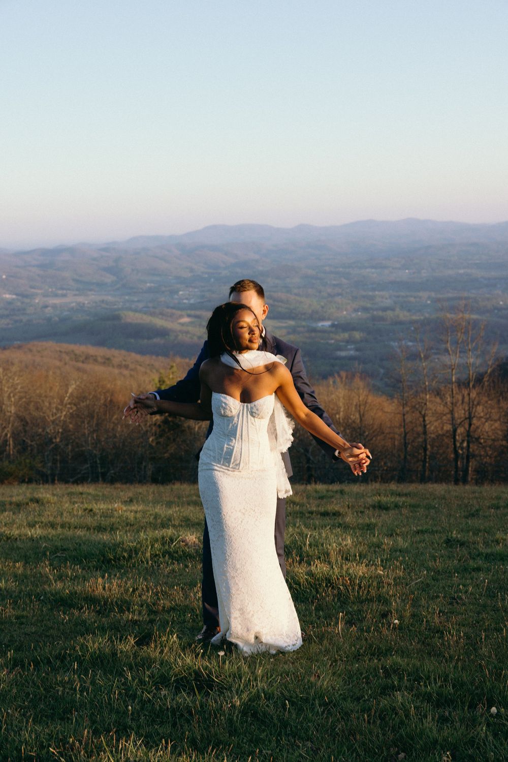 Bride in white strapless gown embraced by groom in navy suit on scenic mountain overlook at golden hour.