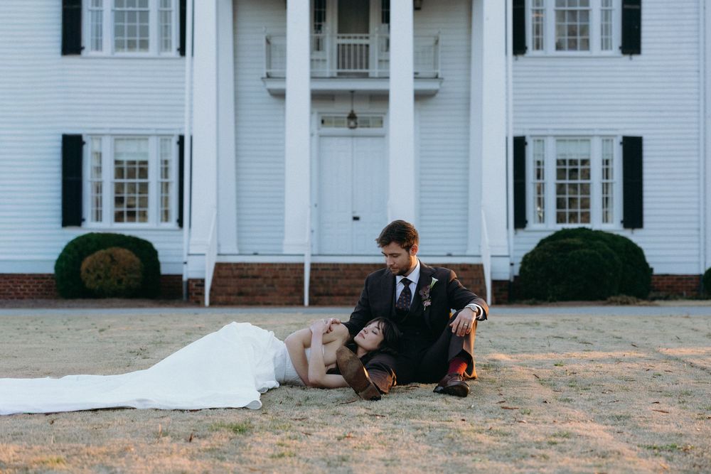 Bride in white gown reclines on grass as groom kneels beside her before a grand white colonial mansion.