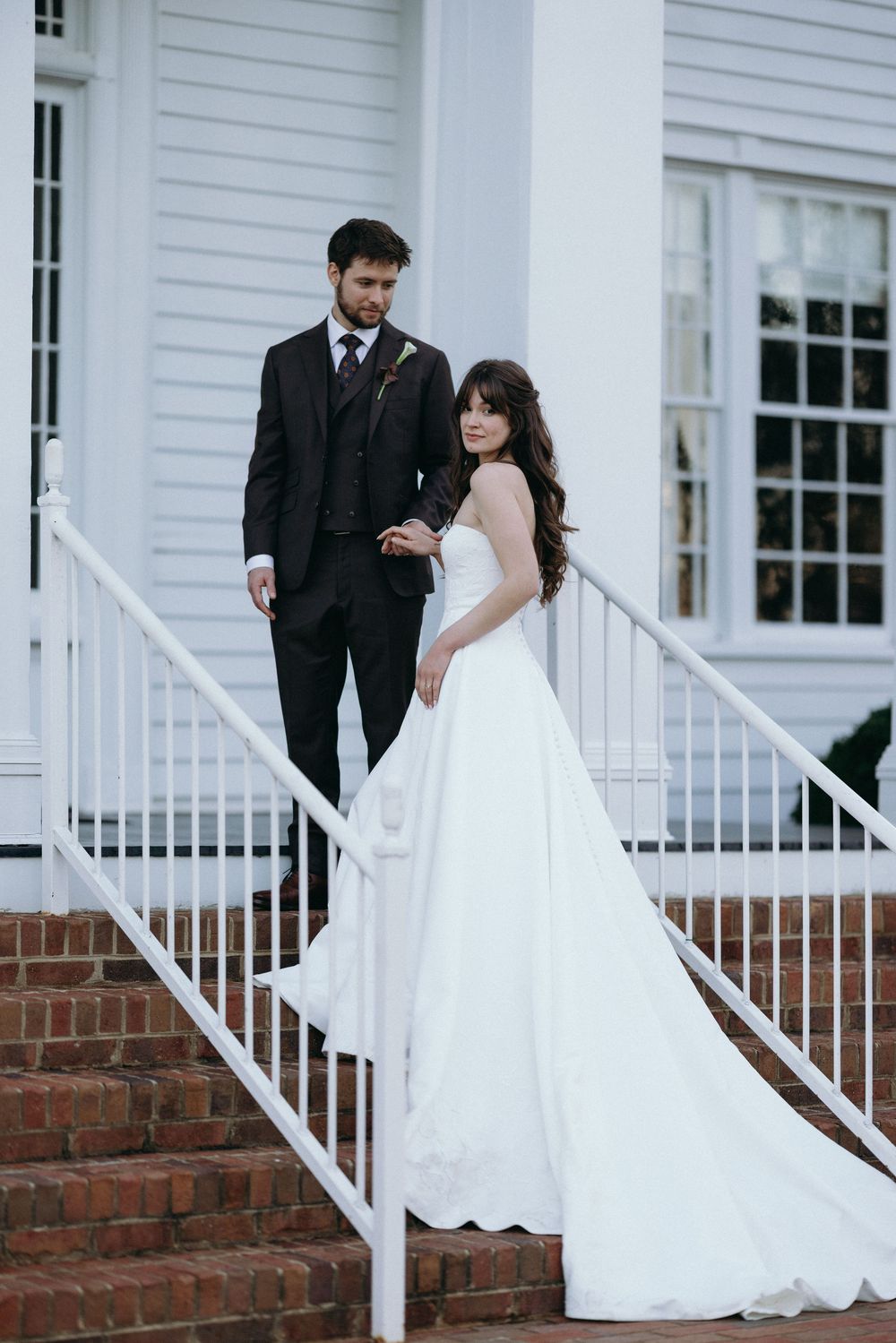 Bride in white ball gown and groom in dark suit pose on brick steps of white colonial building.