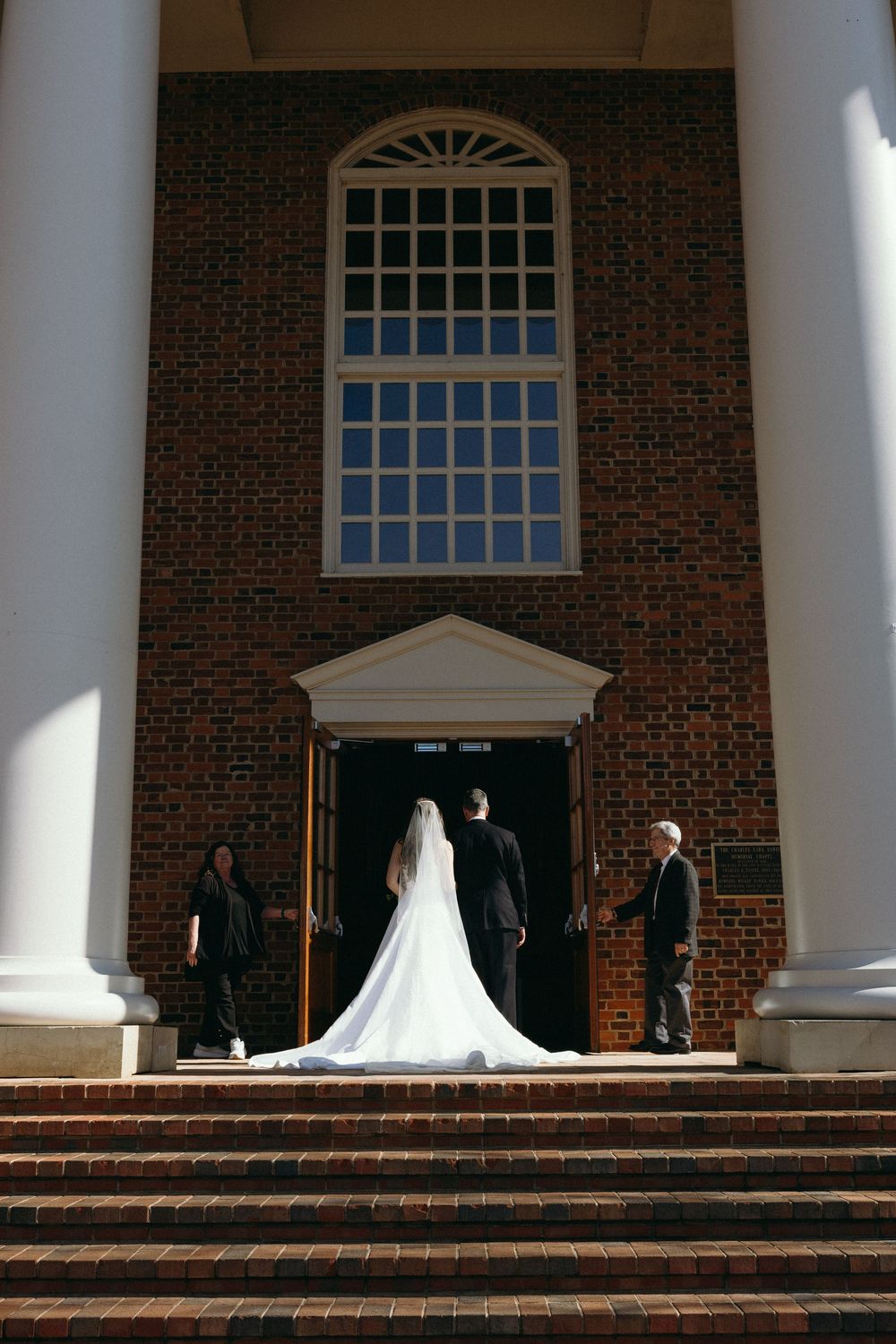 Bride in white gown with groom entering brick church doors, flanked by guests on brick steps with white columns.