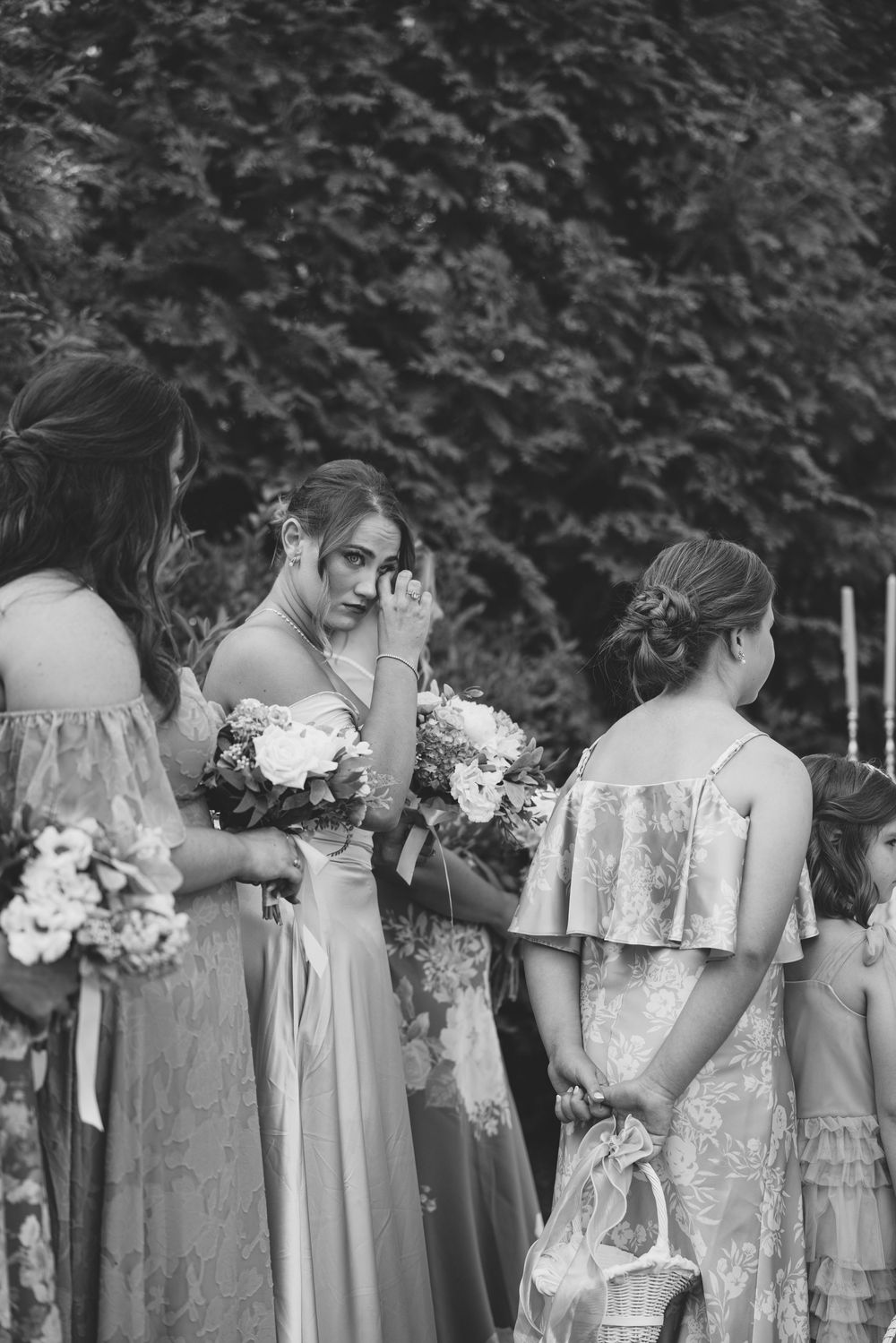 Black and white photo of emotional bridesmaids holding bouquets at a wedding ceremony outdoors.