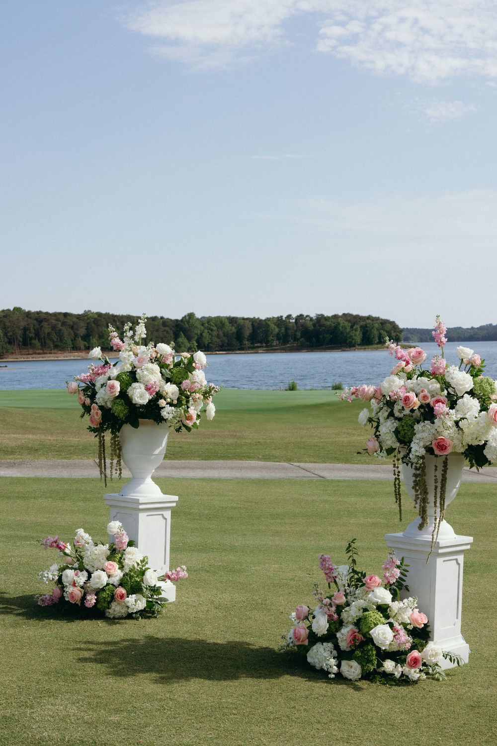 Elegant outdoor wedding ceremony setup with white floral urns on pedestals, pink and white blooms, overlooking a serene lake.