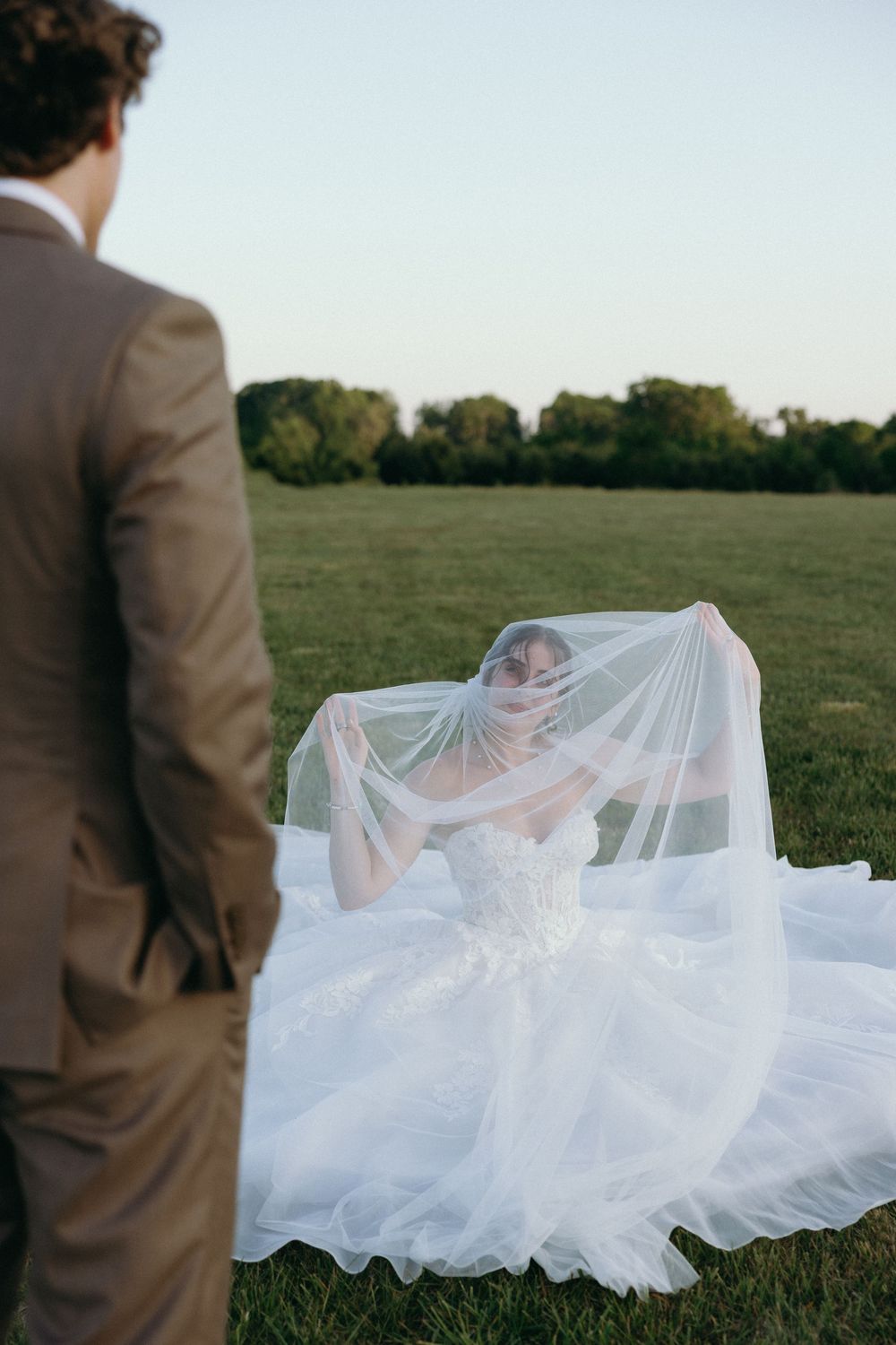 Bride in white ballgown and veil sits on green field as groom in tan suit stands nearby during outdoor wedding.