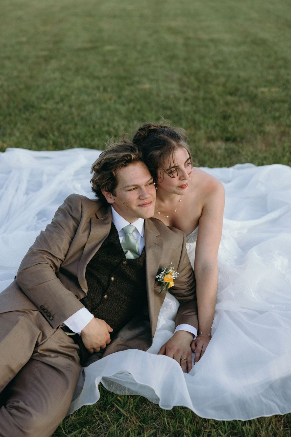 A bride in a white gown and groom in a tan suit sit together on green grass, sharing a romantic wedding moment.