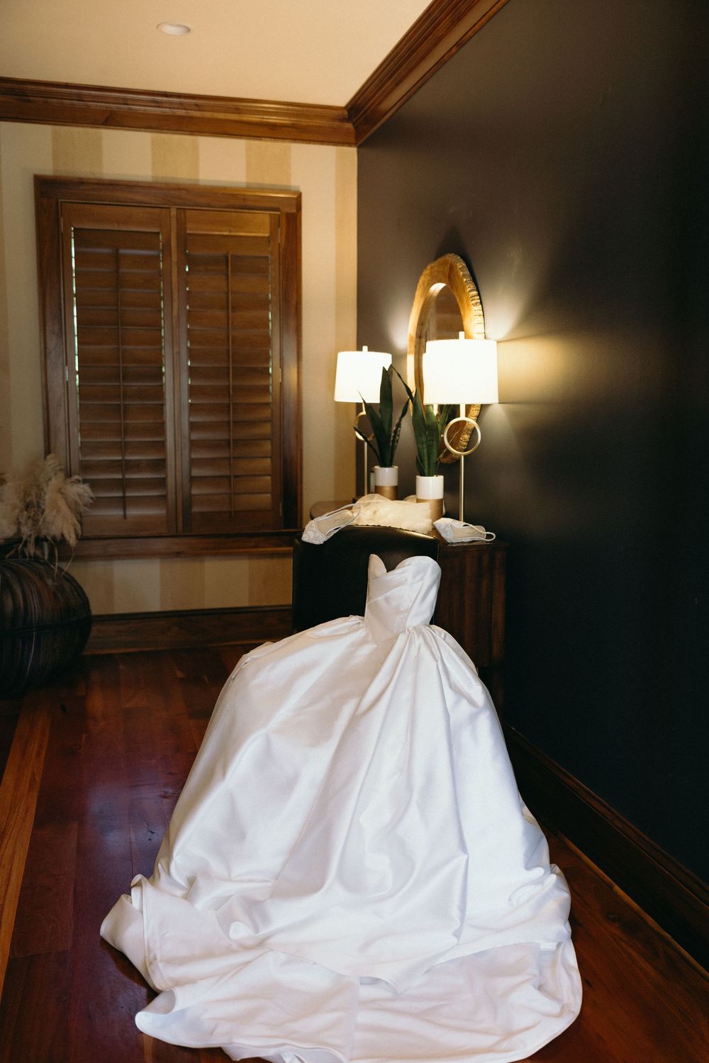 Elegant white ballgown wedding dress displayed on a stand in a dark-walled room with wood floors and warm sconce lighting.