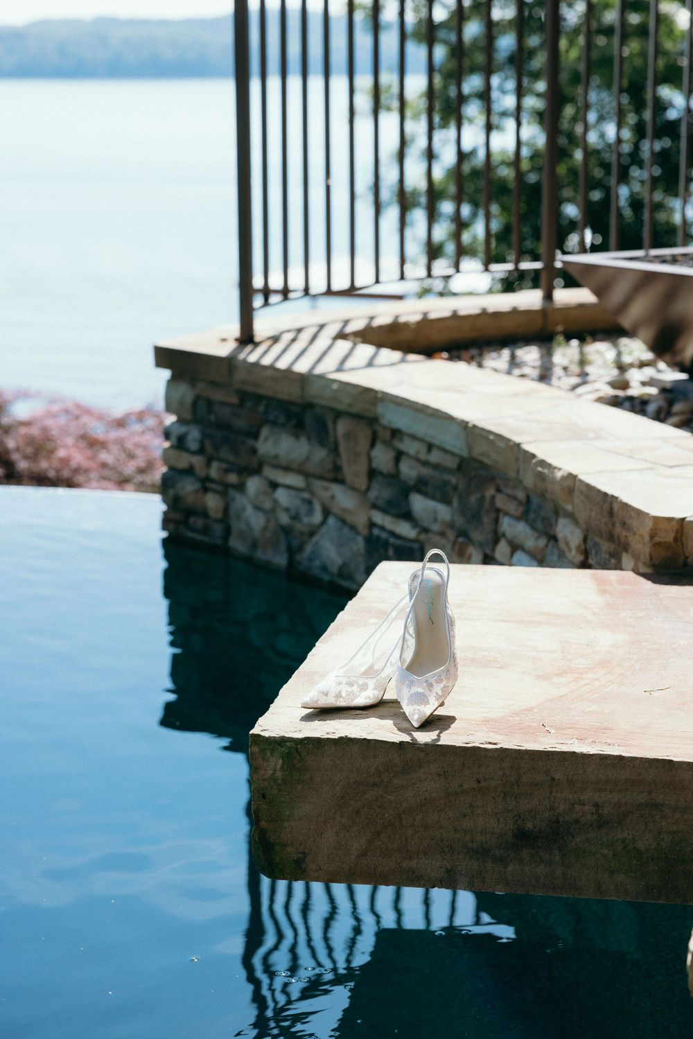 White bridal heels resting on stone edge of infinity pool overlooking serene lake with lush greenery.