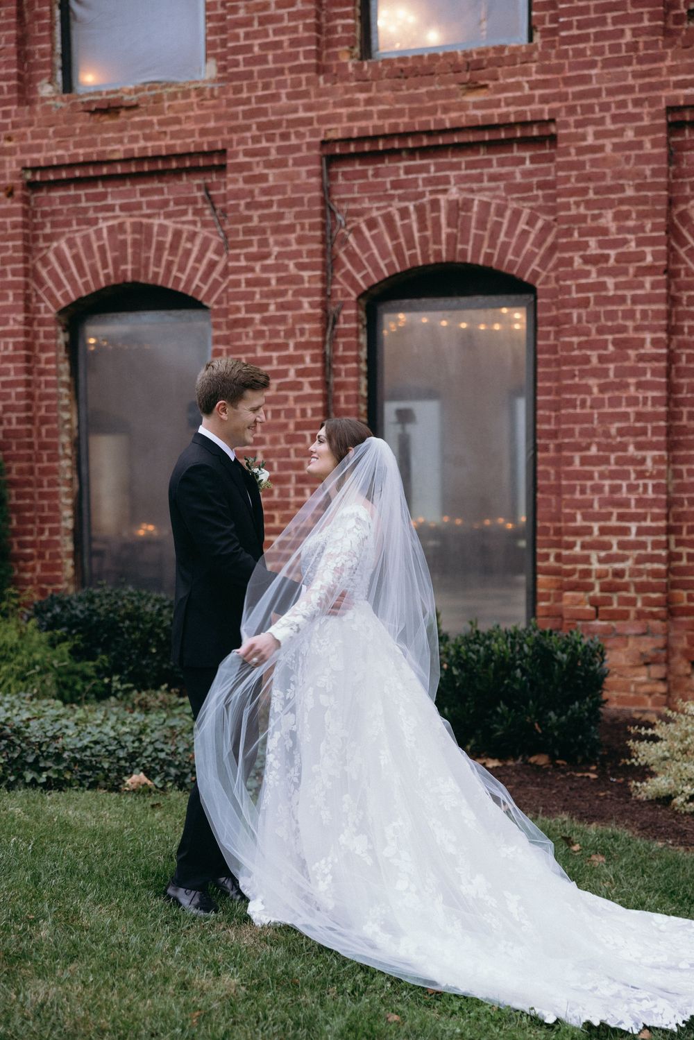 Bride in lace gown with veil and groom in tuxedo walking together outside a red brick building.