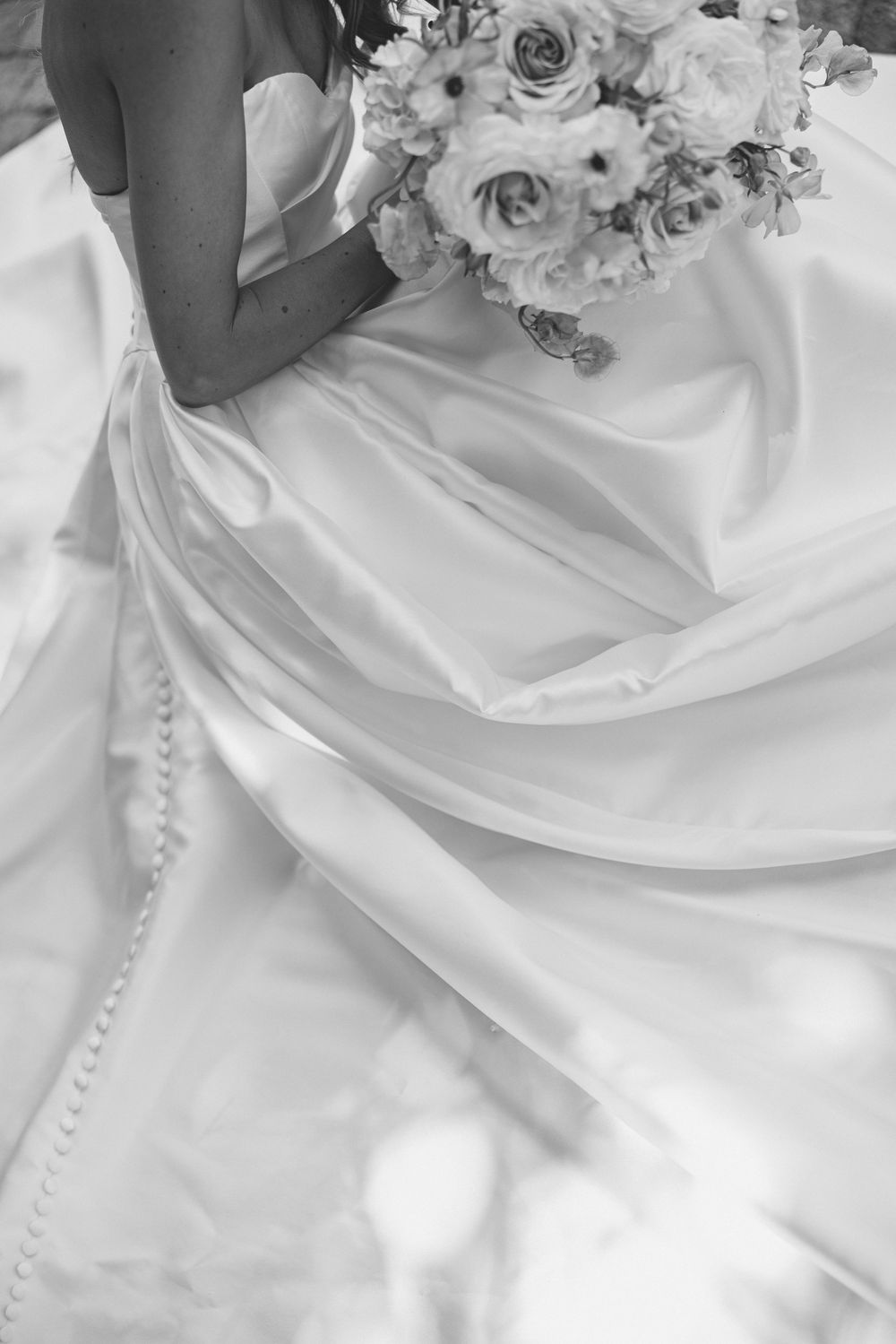 Black and white photo of a bride holding a floral bouquet, showcasing the elegant draping of her white satin wedding gown.
