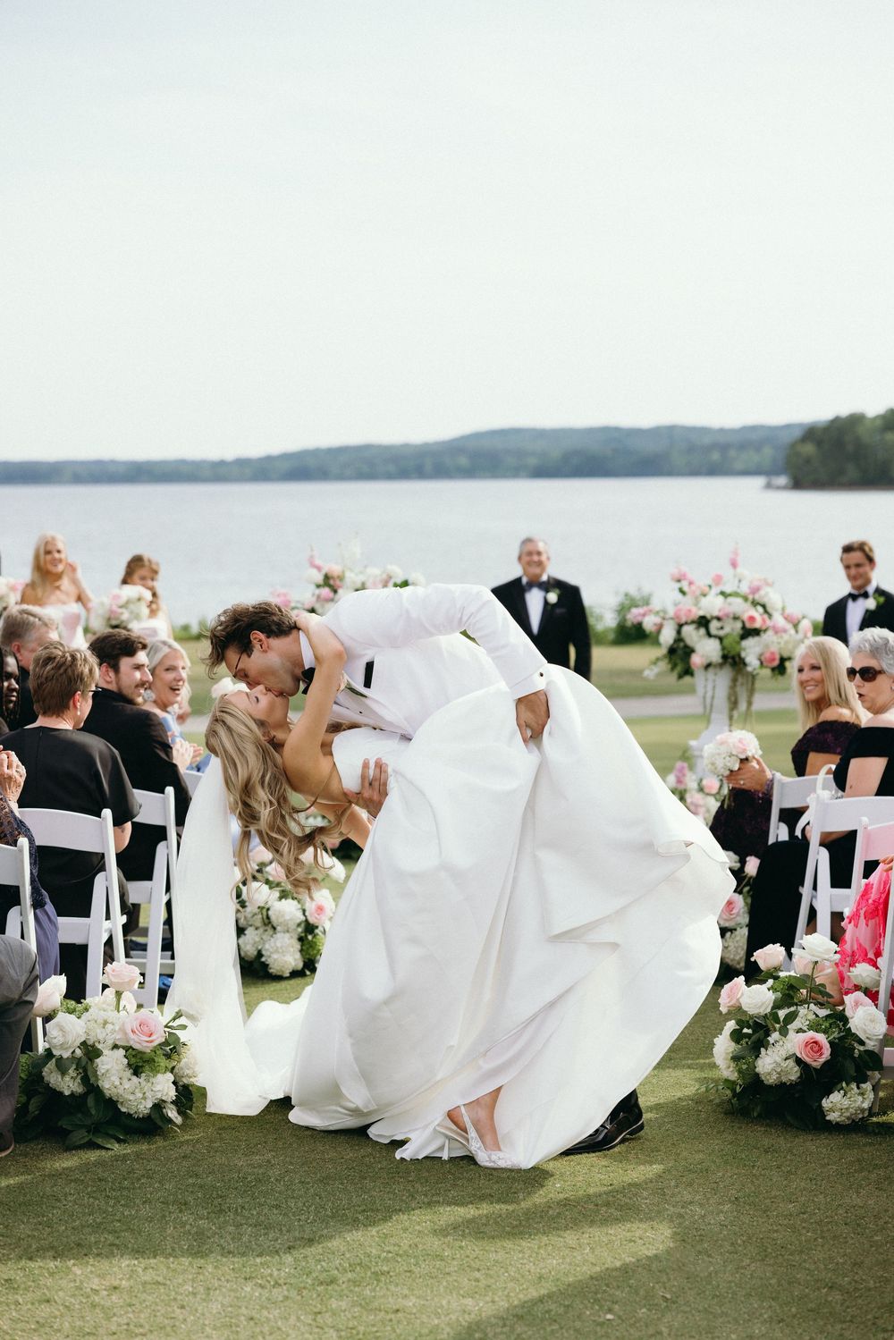 Groom dips bride for a romantic kiss at outdoor lakeside wedding ceremony with floral arrangements and seated guests.