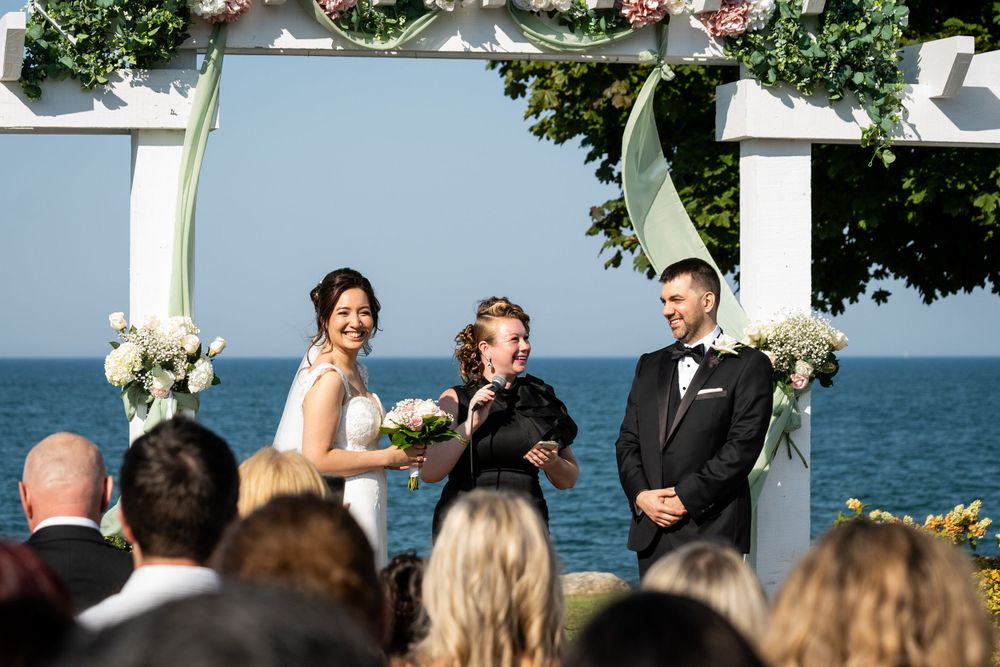 A seaside wedding ceremony with white floral arch decorations and gathered guests.