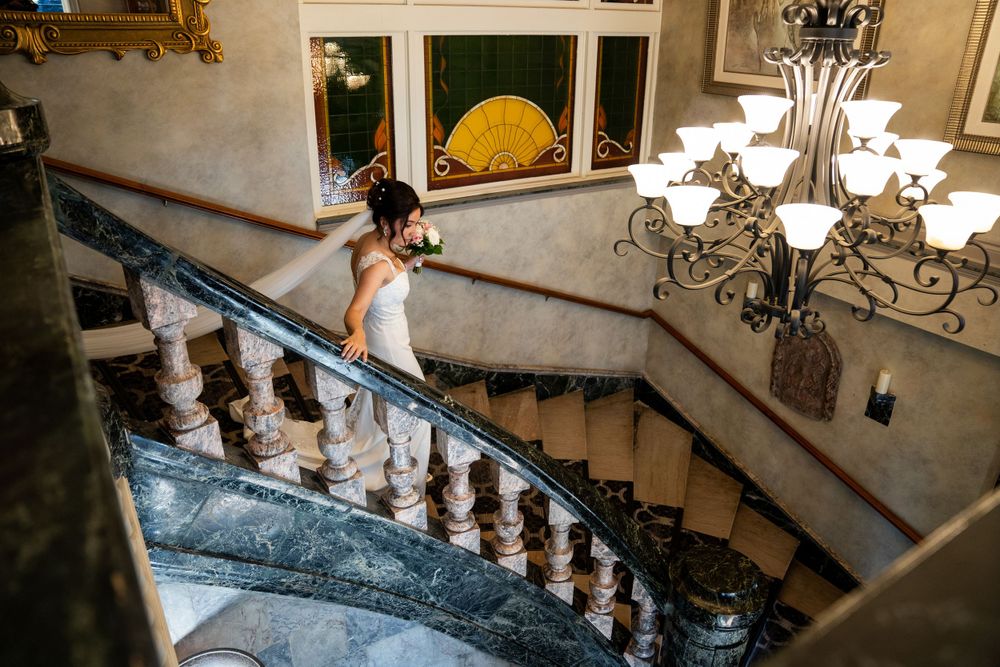 A person in formal wear descending an elegant marble staircase with ornate railing and chandelier.
