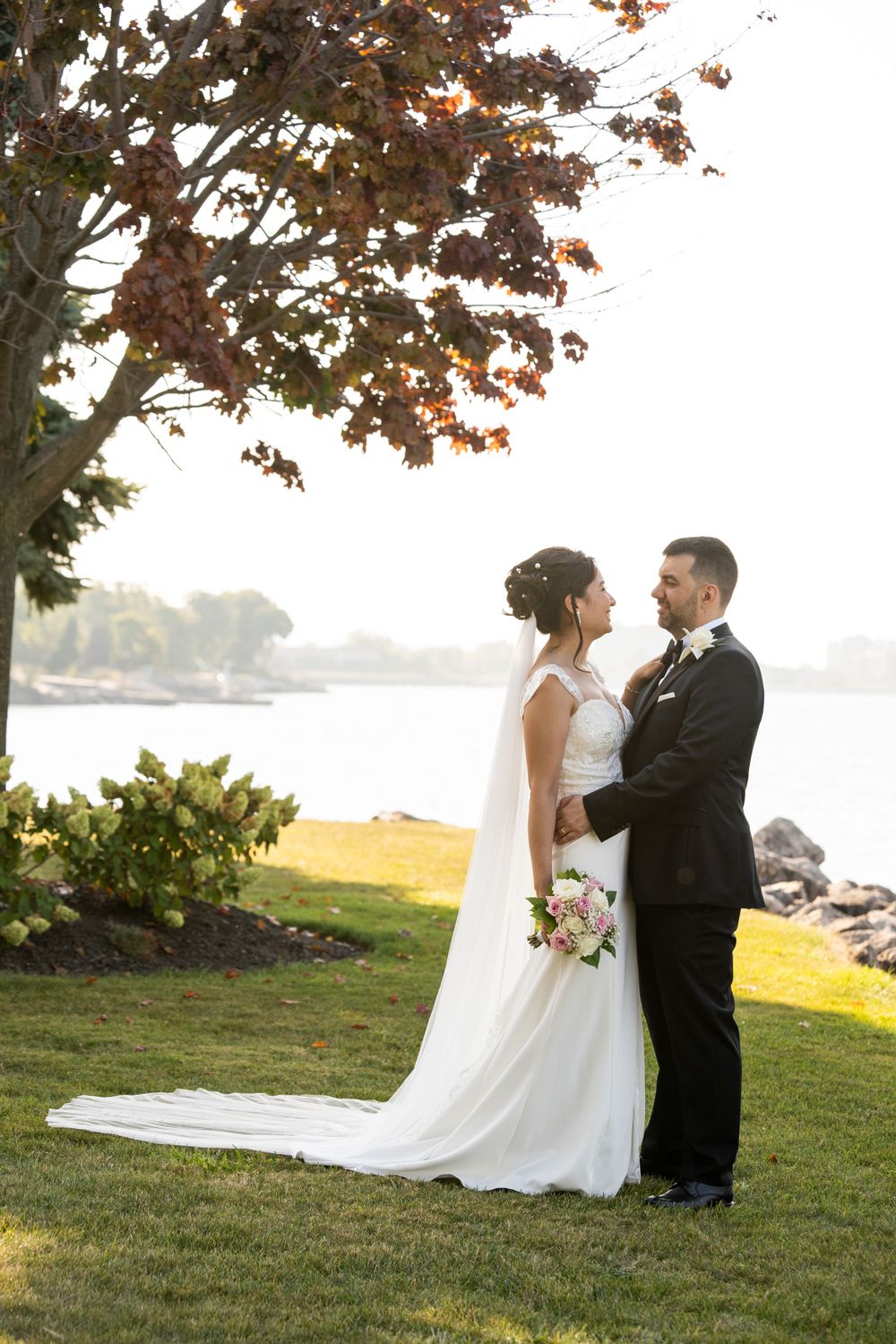A wedding couple poses on a scenic lawn overlooking a waterfront during autumn.