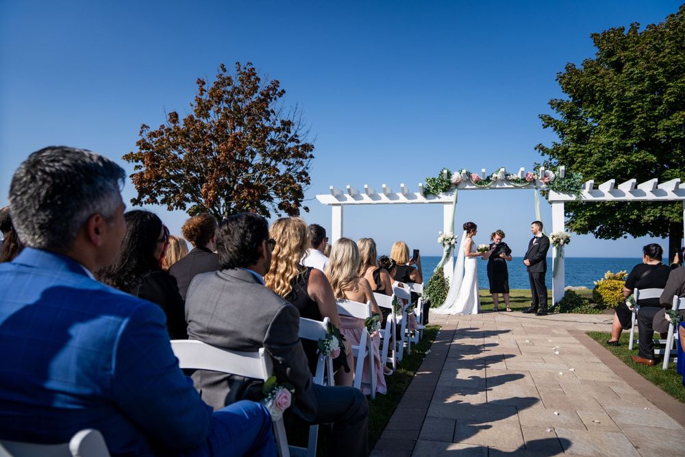 Outdoor waterfront wedding ceremony with white chairs and pergola under blue skies.