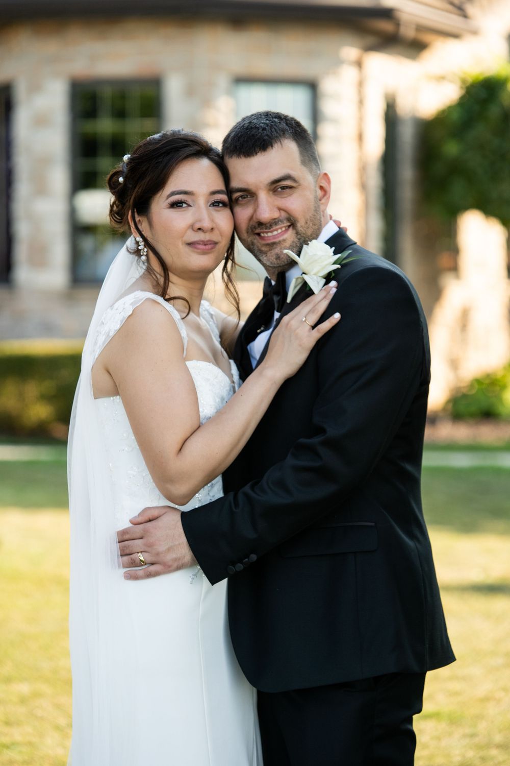 A newlywed couple poses together in formal wedding attire on manicured grounds.