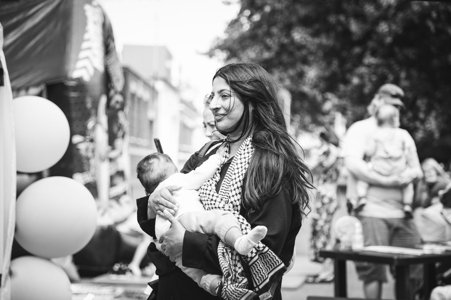 A caregiver holds an infant at an outdoor celebration with balloons.