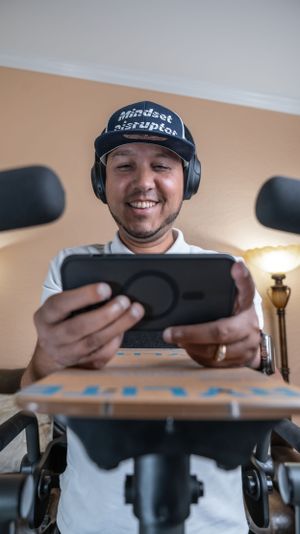 Individual wearing headphones and a baseball cap smiles while looking at handheld device during indoor workout session.