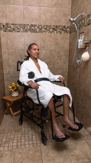 Individual relaxing in a specialized shower chair wearing white robe with sunflowers on side table.