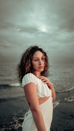 Atmospheric seaside portrait with curly hair and white dress against dramatic cloudy sky.