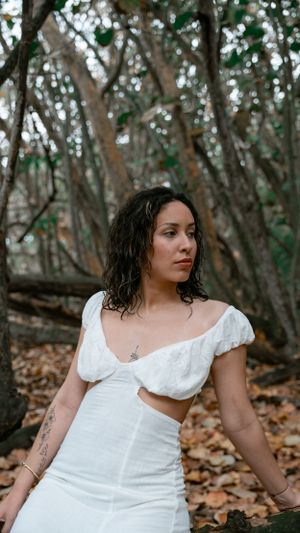 A white crop top and skirt ensemble photographed against a wooded forest backdrop with fallen leaves.
