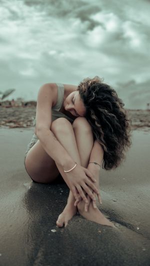 Artistic portrait of person in contemplative pose sitting on wet sand under moody sky.