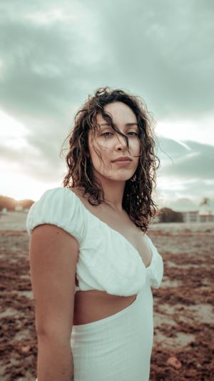 Windswept portrait in white crop dress against stormy sky and rocky terrain.