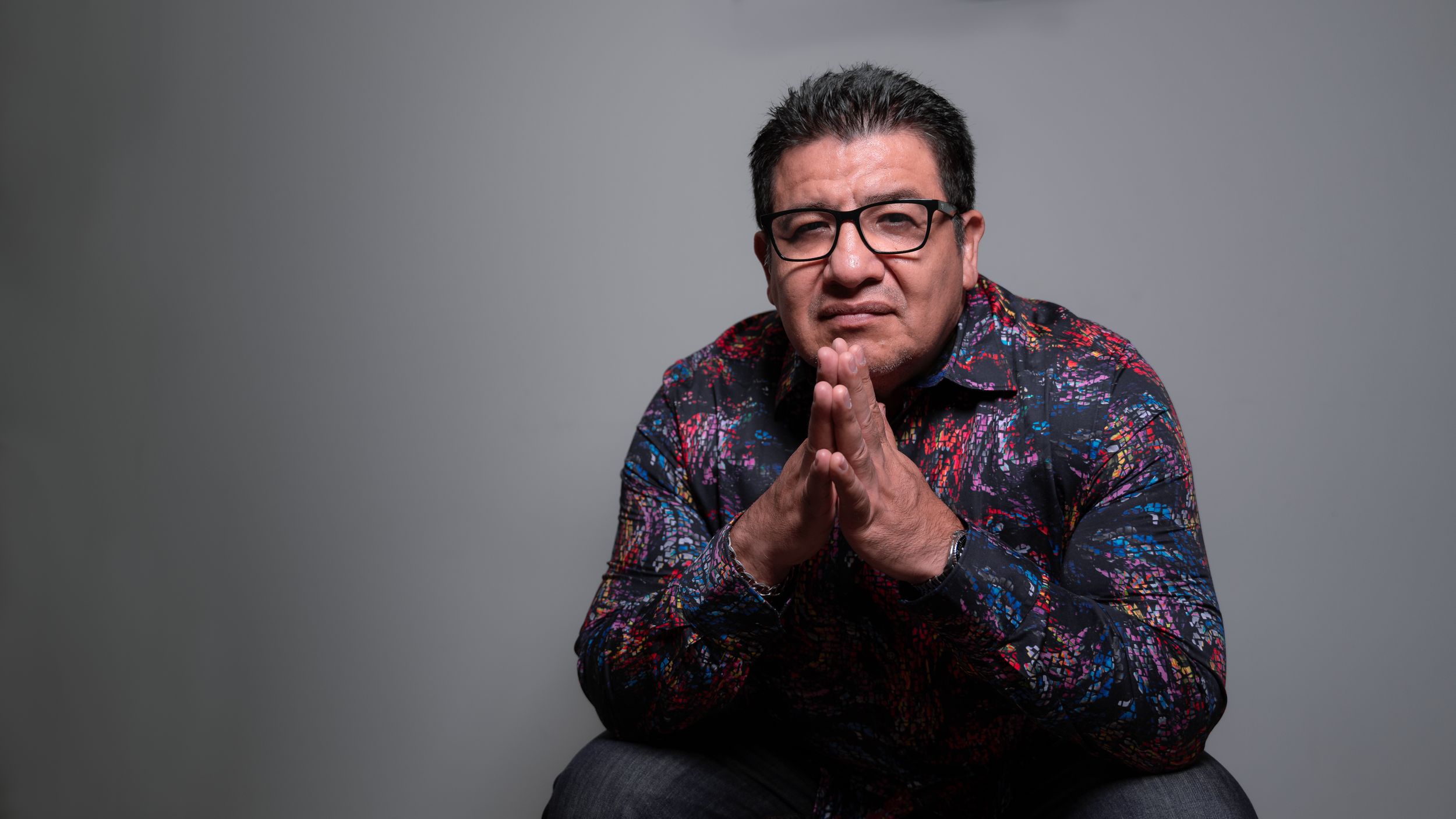 Portrait of a person wearing glasses and a patterned shirt against a gray background.
