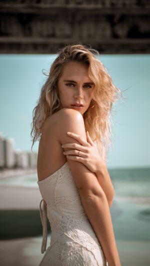 Model in white lace dress poses dramatically against coastal backdrop.