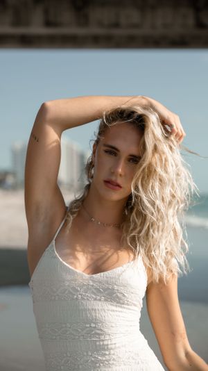 Woman in white tank top poses with hand in blonde hair against beach background.