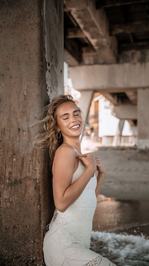 Woman in white dress laughs candidly while leaning against concrete pillar near ocean.