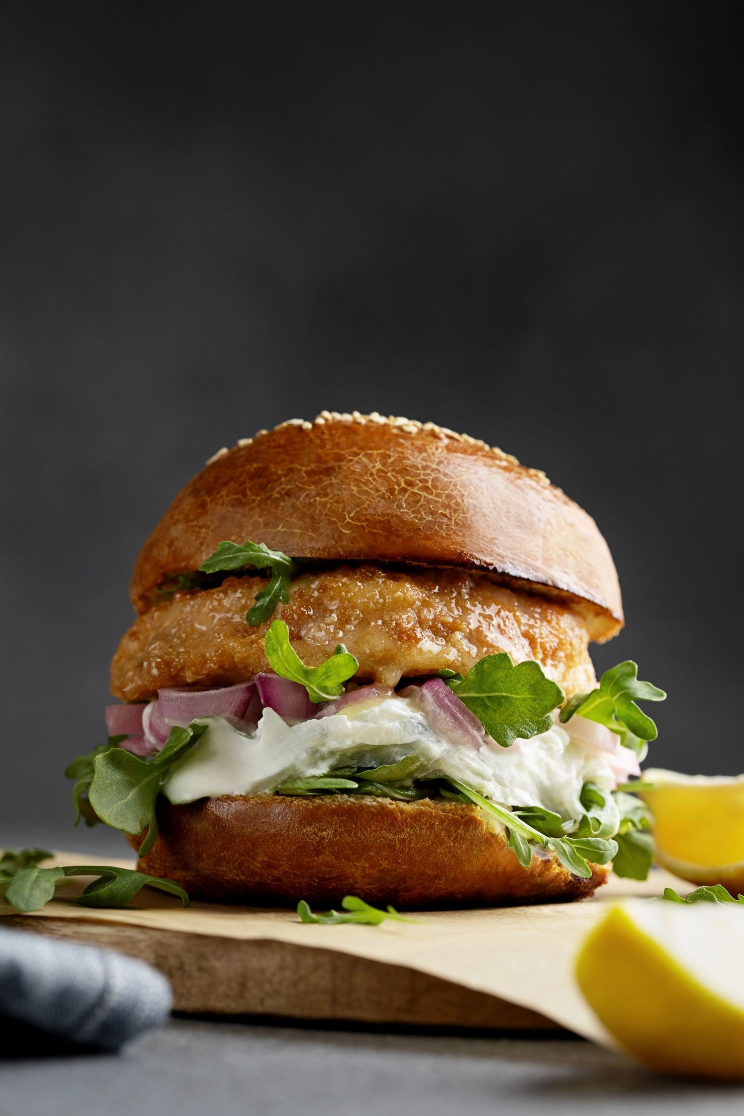 Golden fish burger on brioche bun with arugula, red onion, and tartar sauce, served on wooden board with lemon.