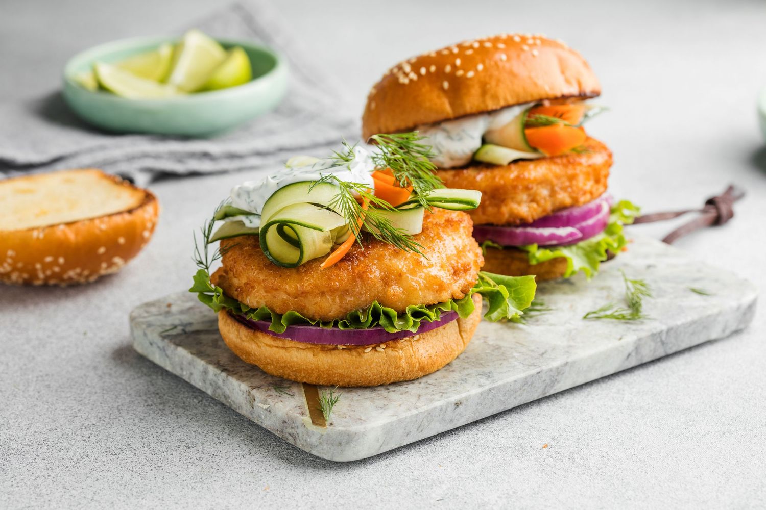 Two crispy fish burgers with cucumber, red onion, lettuce, and microgreens on sesame buns, served on a marble board.