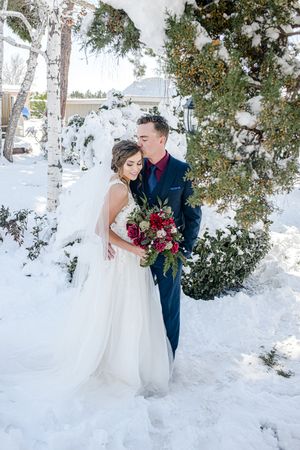 Winter wedding portrait in snow with pine trees and red floral bouquet.