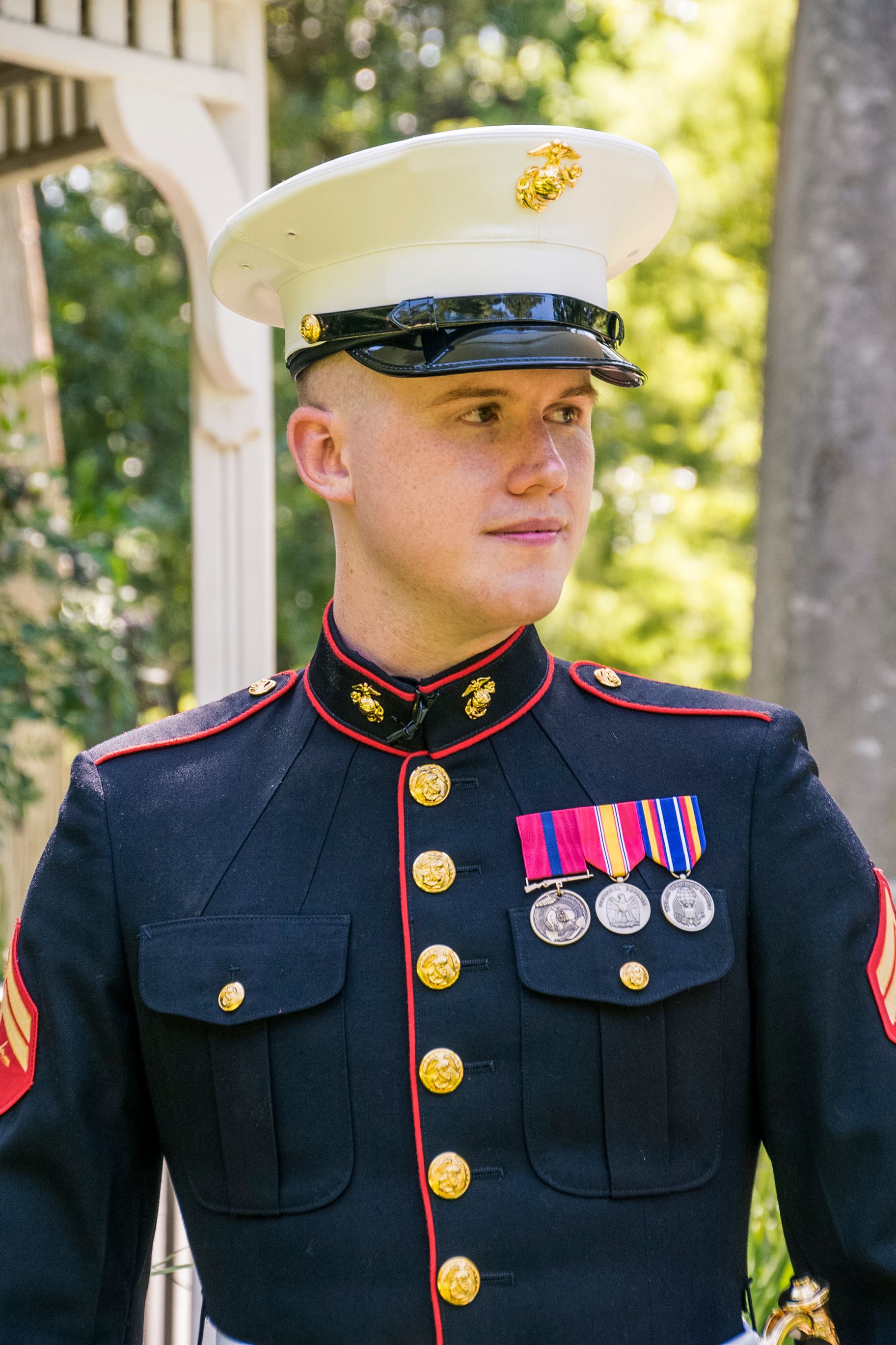 Young US Marine in dress blues uniform with medals and white cap stands outdoors in dappled sunlight.