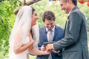 Outdoor wedding ceremony with couple and officiant sharing a joyful moment under trees.