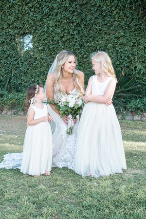 Three people in white dresses sharing a happy moment on grass with ivy wall backdrop.