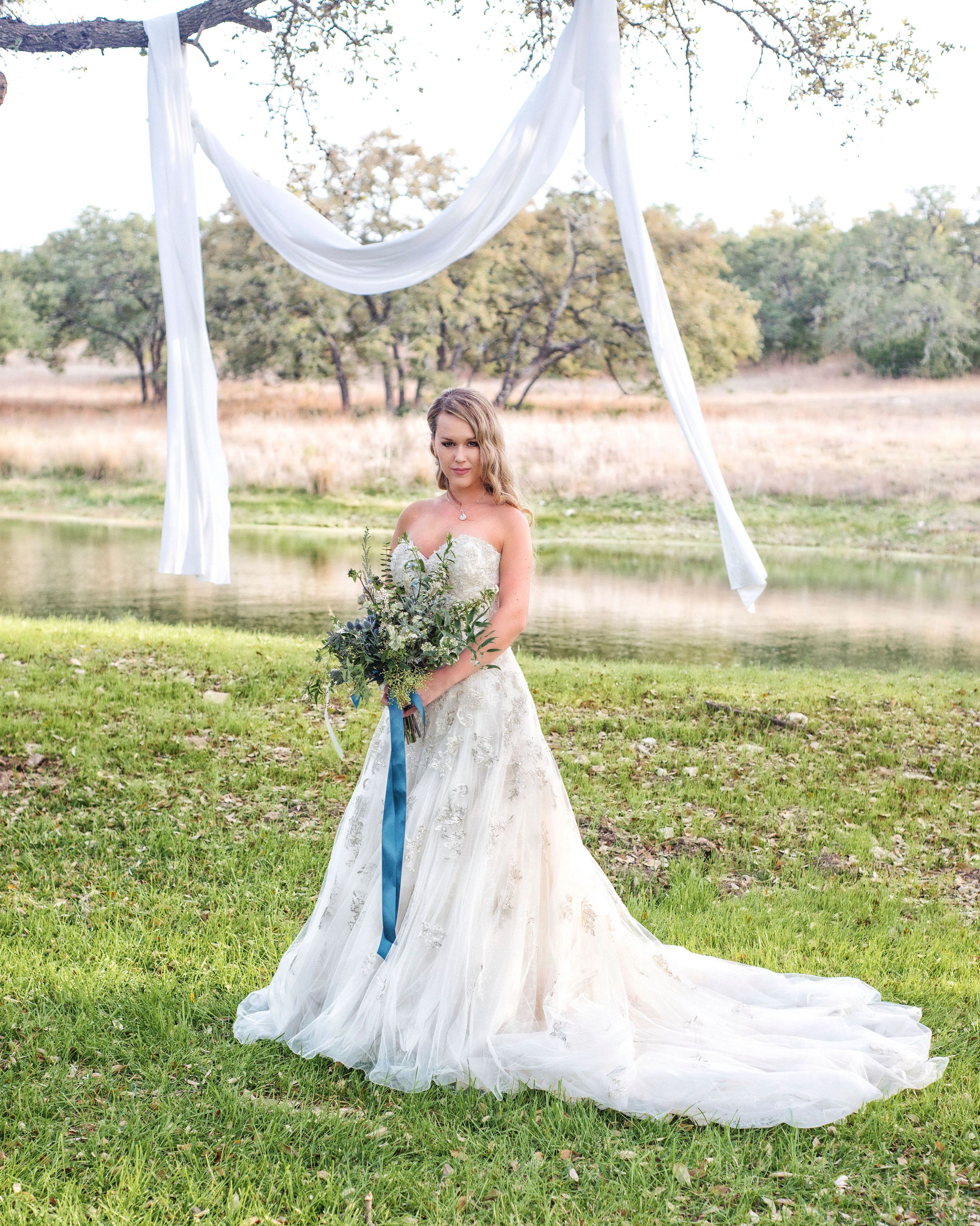 Bride in strapless lace wedding gown holding blue-ribboned bouquet standing under white draped arch by serene pond.