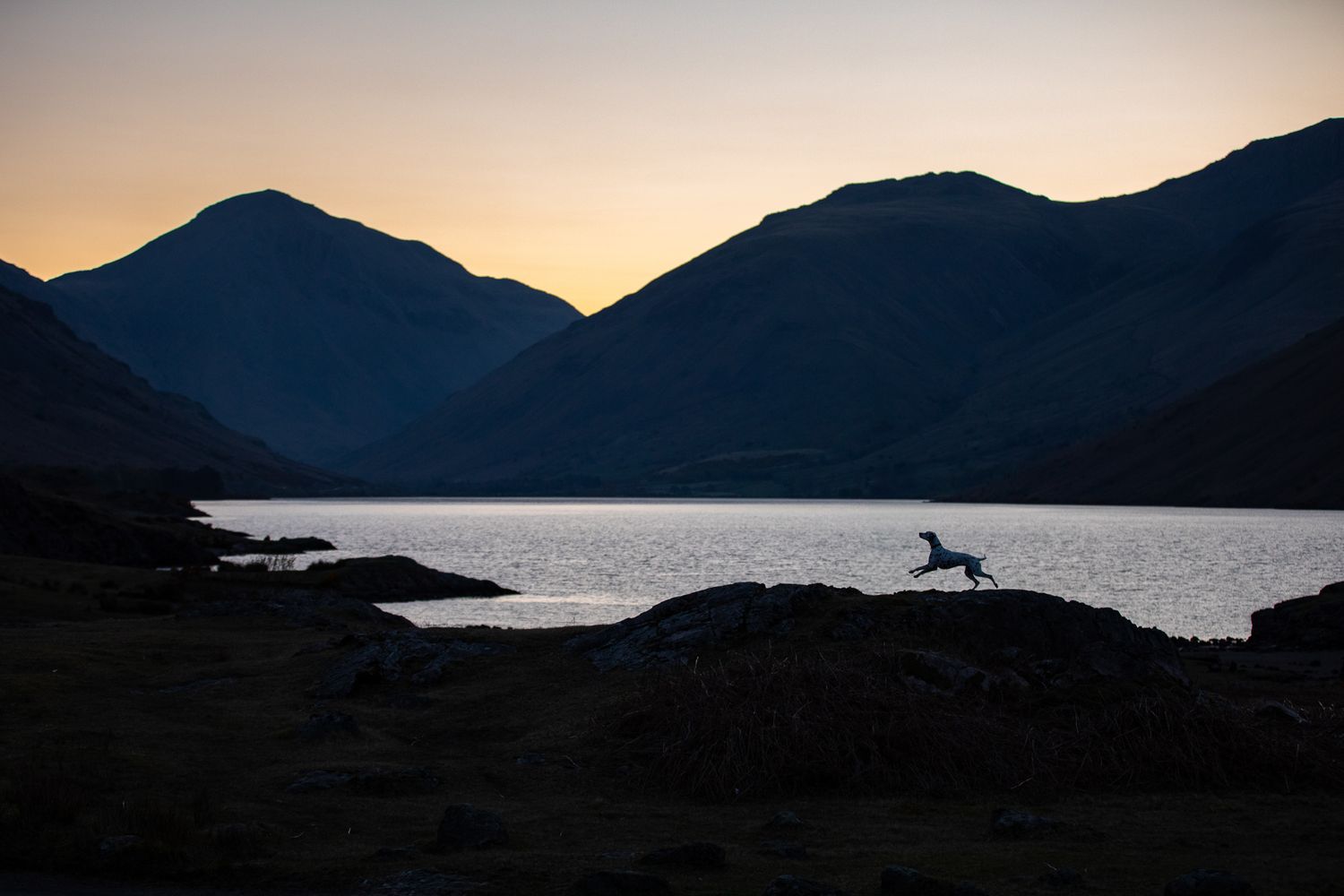 Silhouette of horse and mountains against sunset sky over a serene lake landscape.