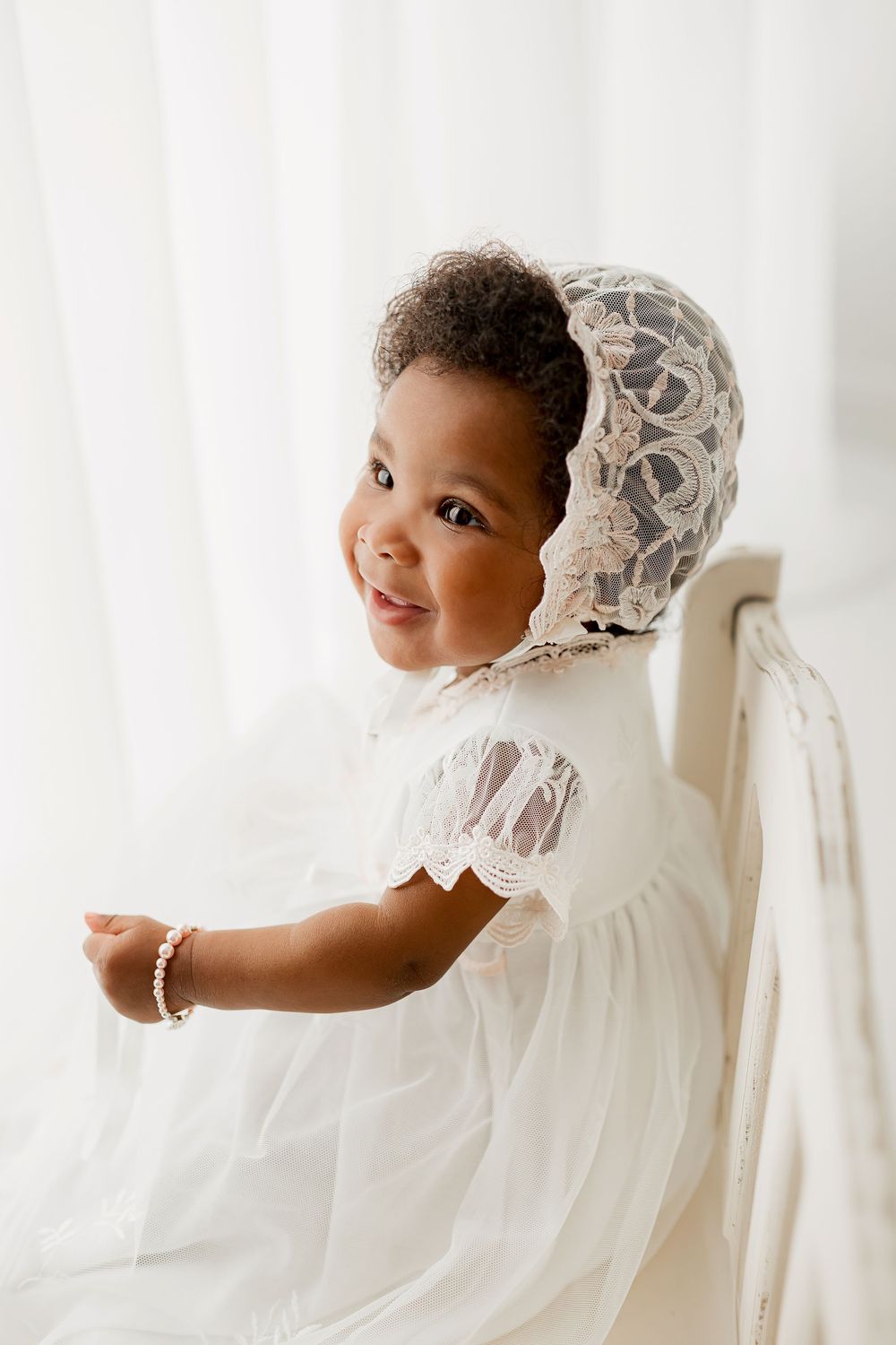 Baby girl in white lace dress and lace bonnet with pearl bracelet standing at vintage bench during milestone portrait session in Summerville