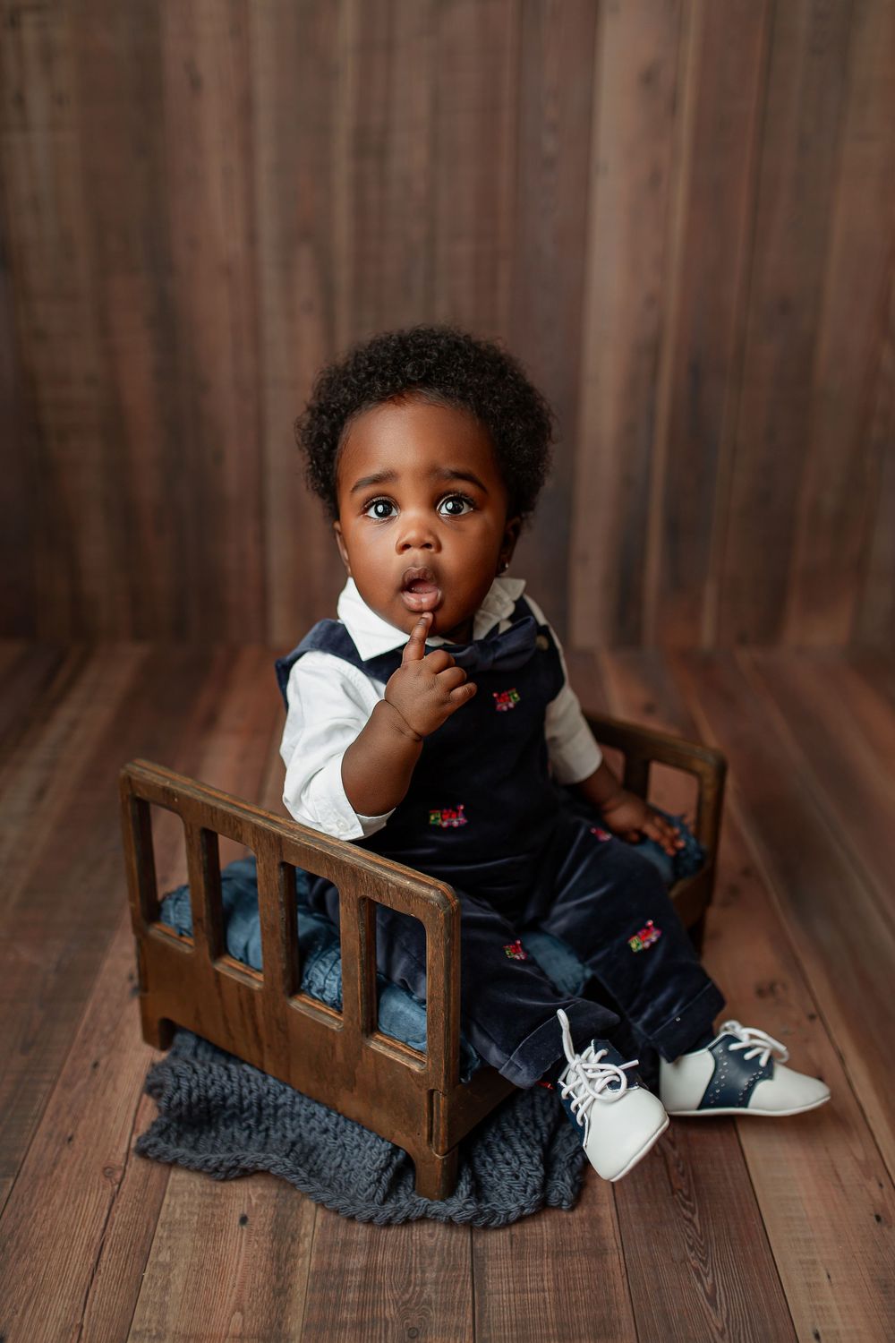 Baby boy in navy velvet outfit with white collar sitting on wooden mini bed with dark wood backdrop during sitter session in Summerville SC