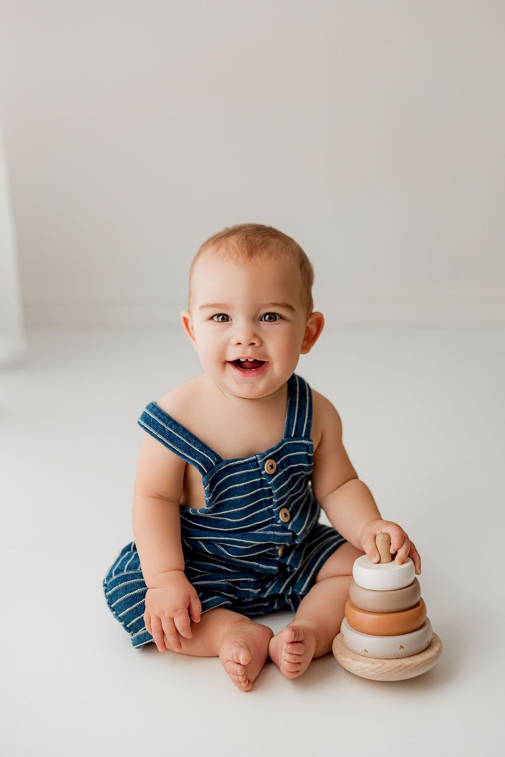 Smiling baby boy in blue striped overalls playing with wooden stacking toy during sitter portrait session at Summerville SC studio