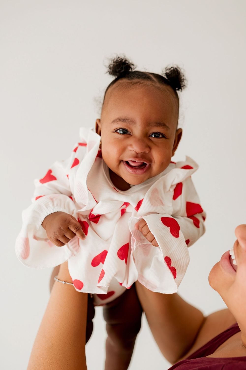 Smiling baby girl with pigtails in white heart print top held up by mom during milestone portrait session in Summerville SC