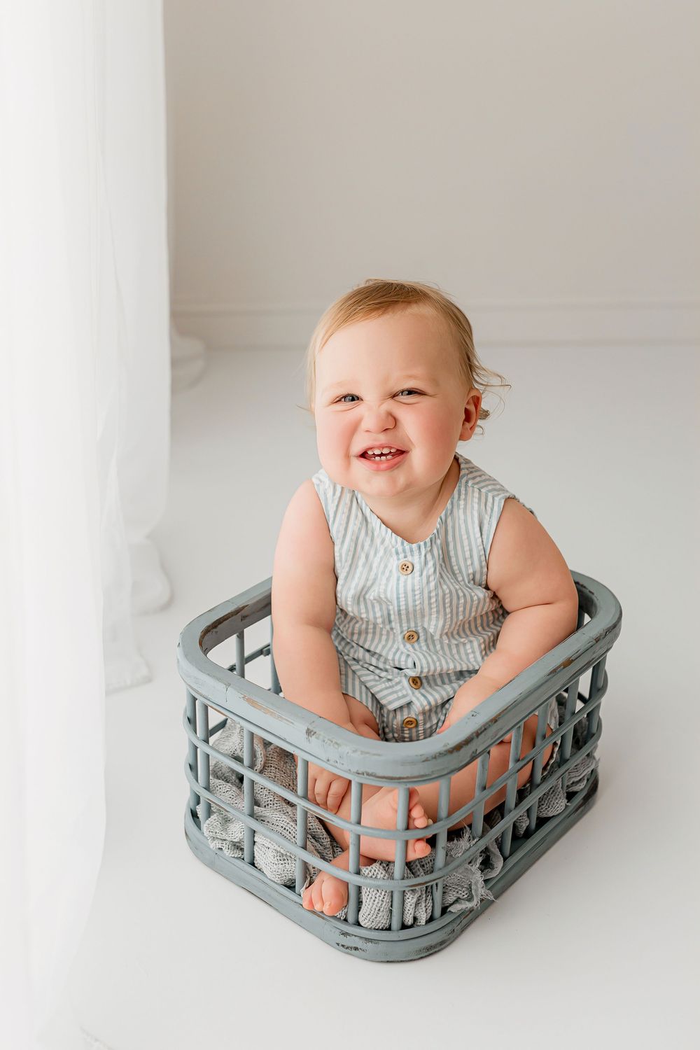 Smiling baby in blue striped seersucker romper sitting in vintage blue painted crate during sitter portrait in Summerville SC studio
