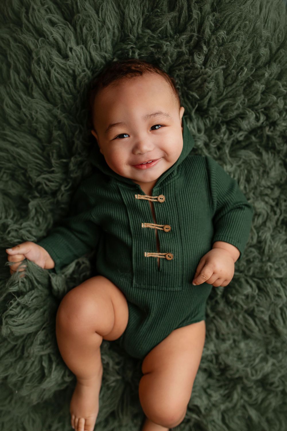 Smiling baby boy in green hooded romper lying on green fur blanket during milestone session at Flowertown Photography in Summerville SC