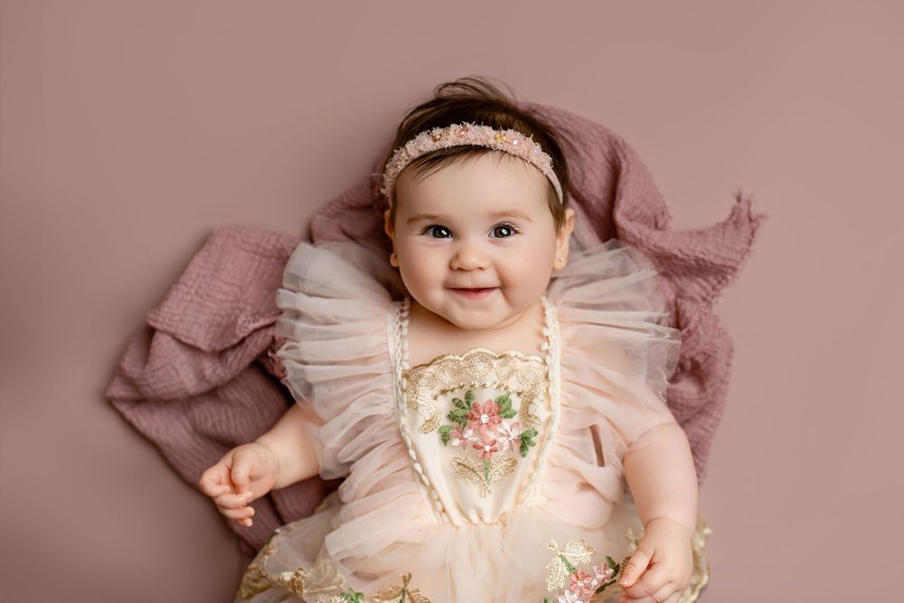 Baby girl in pink embroidered tulle dress with floral headband smiling on mauve backdrop during milestone session in Summerville SC